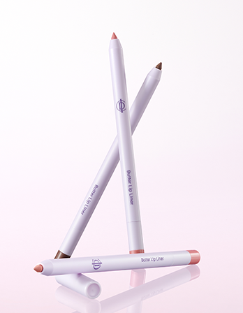 Three lip liner pencils arranged in a crossed and horizontal layout on a reflective light pink and white background. The pencils are white with peach-colored tips and brown tips, and the brand logo is visible on each.