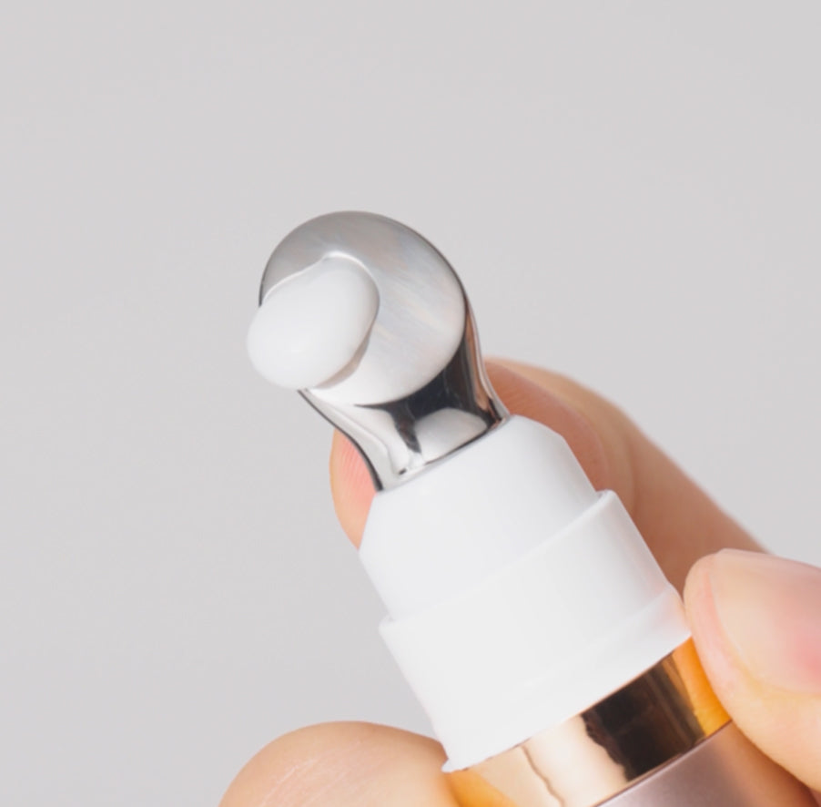 A close-up of a hand holding a cosmetic pump bottle with a white silicone droplet on a metallic silver nozzle, set against a soft light gray background.