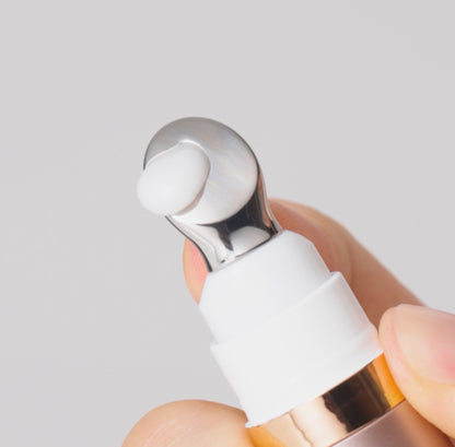 A close-up of a hand holding a cosmetic pump bottle with a white silicone droplet on a metallic silver nozzle, set against a soft light gray background.
